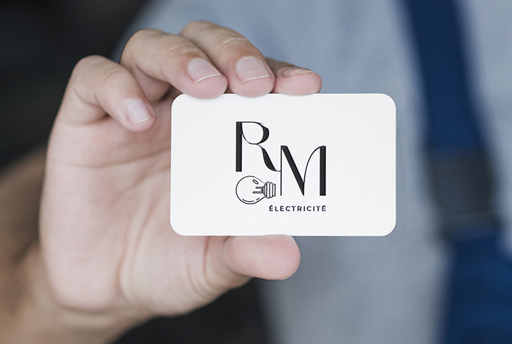 Carte de visite blanche tenue entre deux doigts, avec les initiales RM en noir stylisé, une icône d’ampoule et la mention « ÉLECTRICITÉ ».