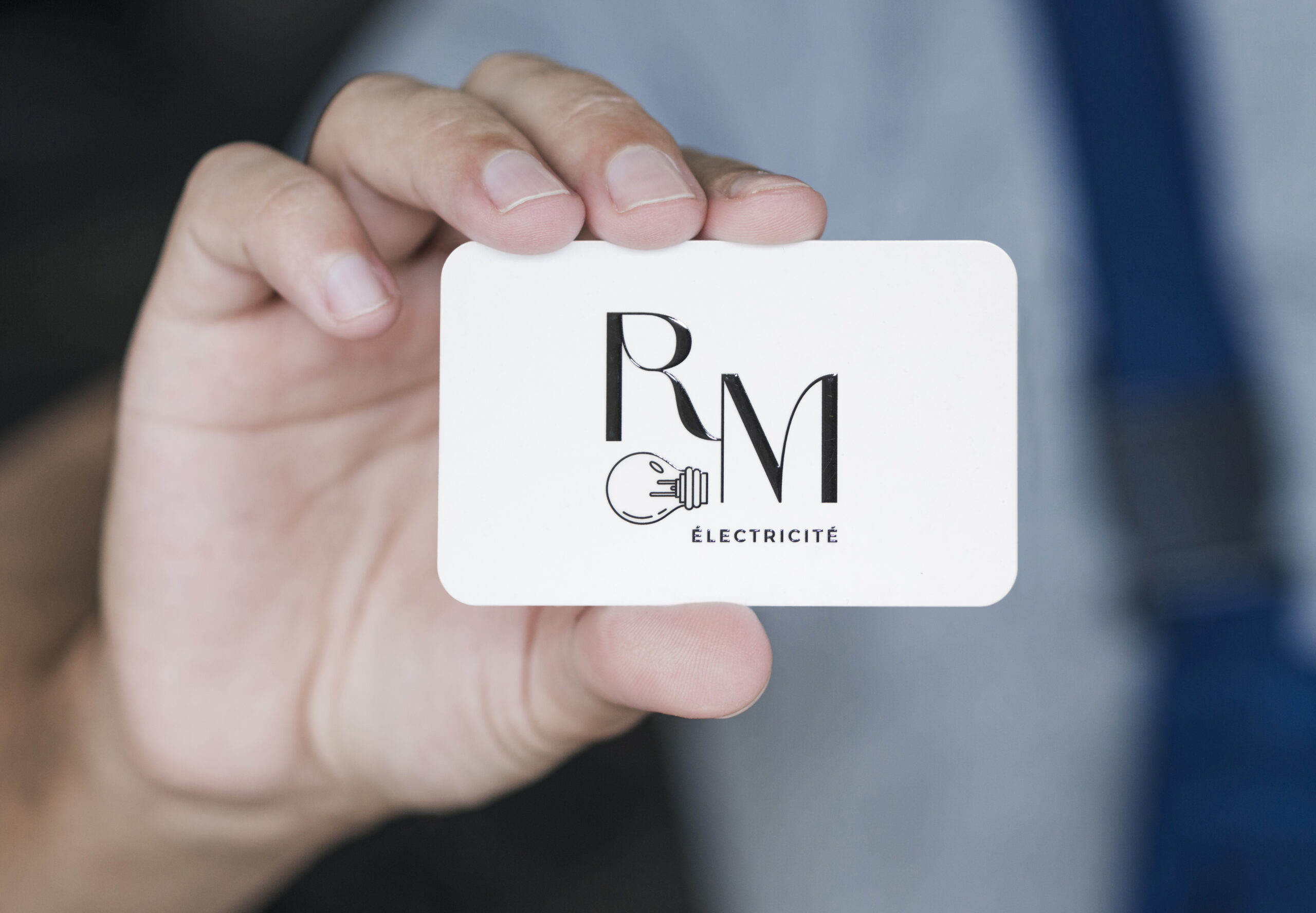 Carte de visite blanche tenue entre deux doigts, avec les initiales RM en noir stylisé, une icône d’ampoule et la mention « ÉLECTRICITÉ ».