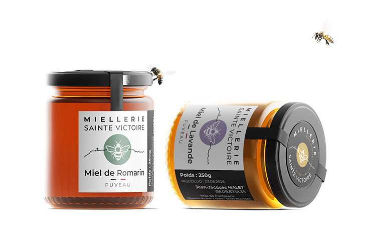 Visuel de deux pots de miel de la Miellerie Sainte Victoire : un pot de miel de romarin en position verticale, l'autre de miel de lavande en position allongée. Étiquettes élégantes avec logo d’abeille stylisée et lignes évoquant le massif de la Sainte-Victoire. Palette sobre (noir, blanc, pastel) et capsules noires. Deux abeilles en vol complètent la scène. Design moderne valorisant l’artisanat et le terroir de Fuveau (Provence).