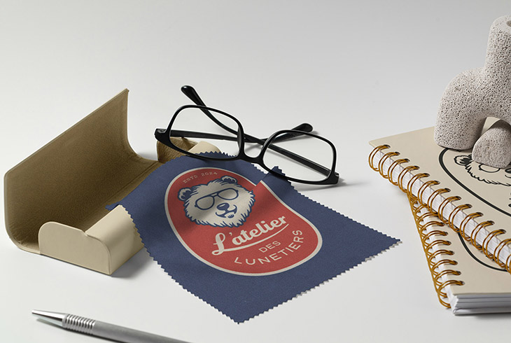 Monture de lunettes noire posée sur un chiffon de nettoyage bleu avec le logo de L’atelier des Lunetiers, accompagné d’un étui ouvert, d’un stylo, et de carnets à spirale personnalisés avec le même logo. L’ensemble est présenté sur un fond blanc, dans une composition soignée évoquant l’univers de la papeterie et des accessoires optiques.